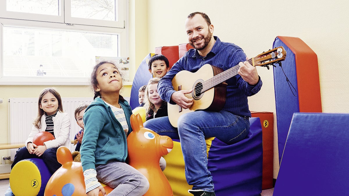 Porträt des Kindergartenpädagogen Daniel Granögger. Er sitzt umringt von Kindern im Kindergarten-Gruppenraum und spielt Gitarre