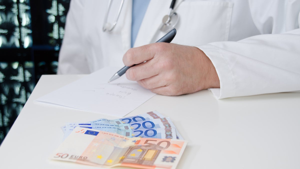 Ein Tisch auf dem im Vordergrund Geld liegt, im Hintergrund ist ein Arzt zu sehen