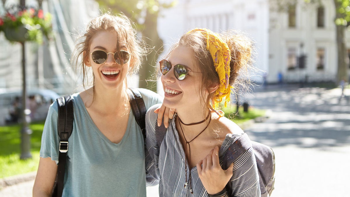 Outdoor portrait of two female comrades walking on street laughing while talking with each other enjoying summer vacations. Two teenagers in sunglasses holding rusksacks on backs going on picnic