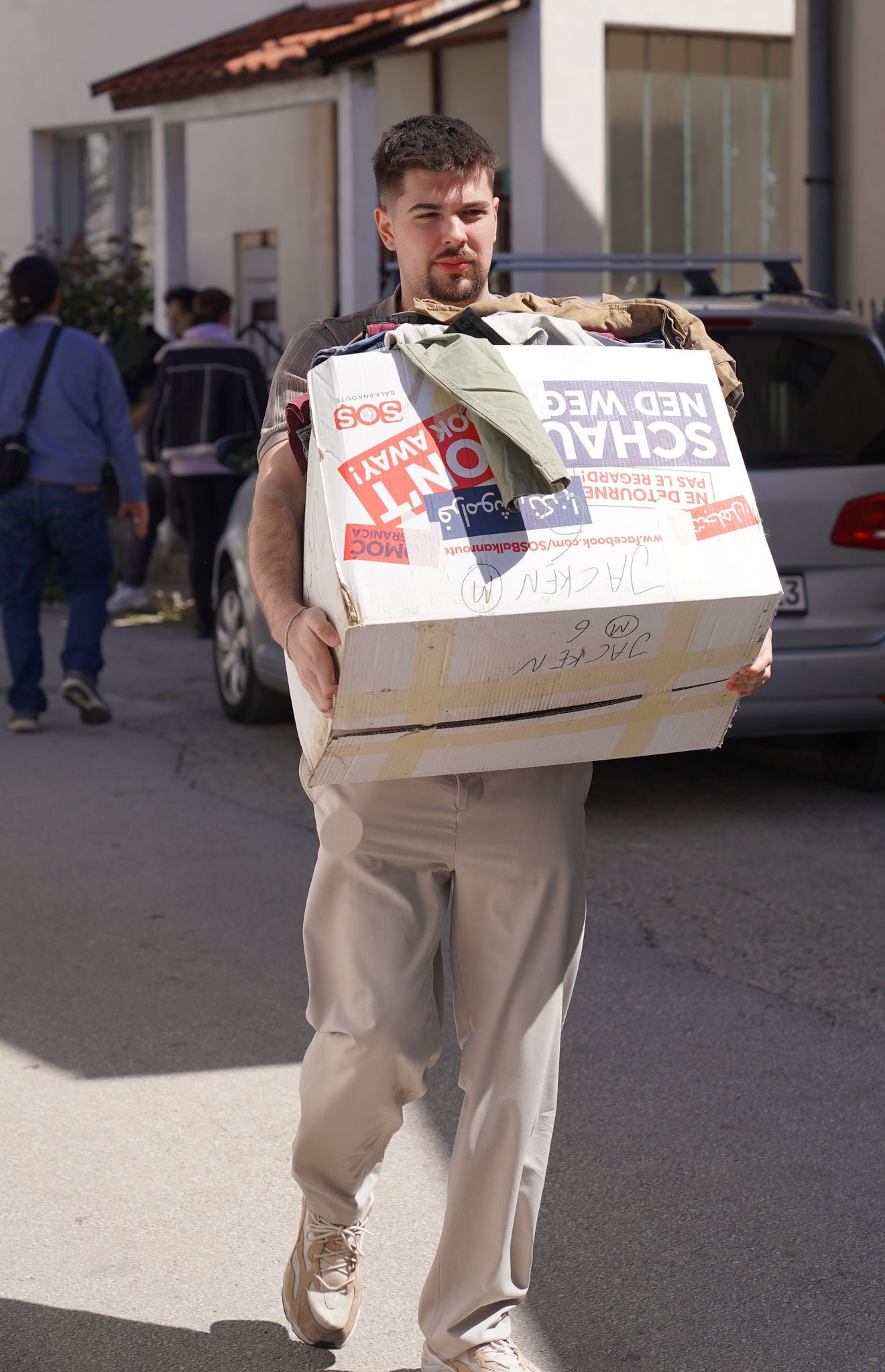 Ein junger Mann transportiert einen großen Karton mit Kleidung vor einem Wohnhaus. Die Szene vermittelt einen Umzug oder die Abgabe von Kleiderspenden.