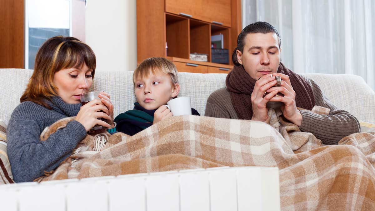 Familie sitzt mit dicken Decken und Schalls im Wohnzimmer und wärmt die Hände an Teetassen.