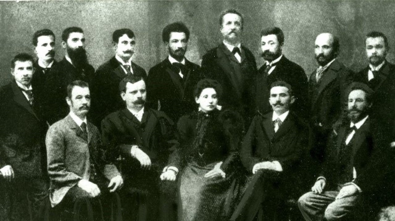 Eine Gruppe von Männern mit beeindruckenden Bärten und vorne sitzend Anna Boschek mit langem Kleid.