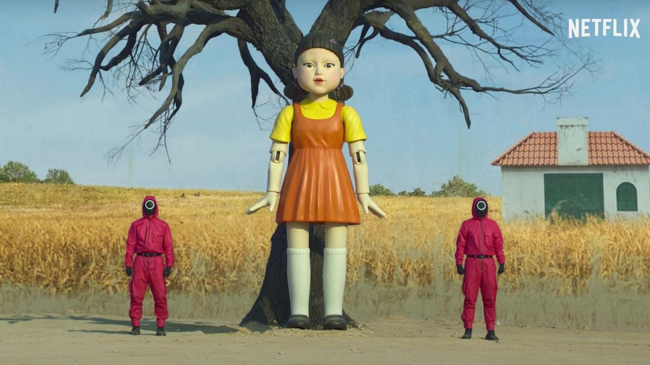 Screenshot der Netflix Serie "Squid Games". Ein Roboter Mädchen steht vor einem Baum und künstlichem Hintergrund, rechts und links von ihr stehen Wachen in dunkel rosa Anzügen und schwarzen Masken.