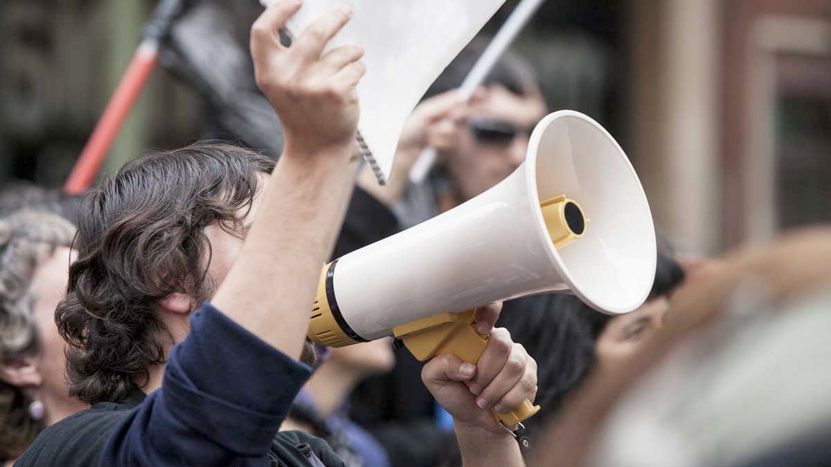 Foto von einem Protest. Im Vordergrund ein Mann mit Megafon der ein Schild hochhält.