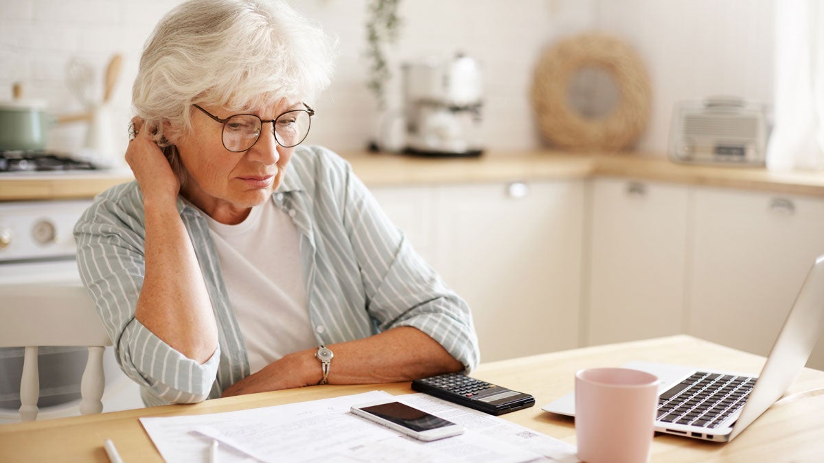 Pensionistin sitzt ratlos am Küchentisch, vor ihr liegen Rechnungen, ein Taschenrechner und ein Handy