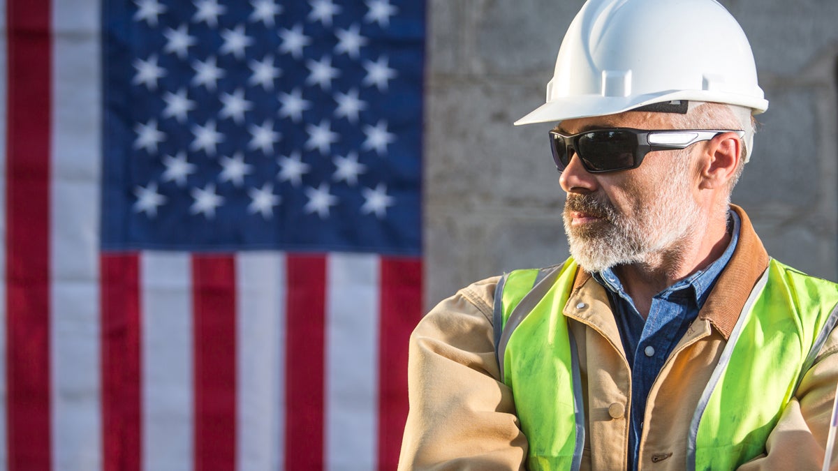Bauarbeiter mit Helm, Schutzbrille und Weste vor einer USA-Flagge