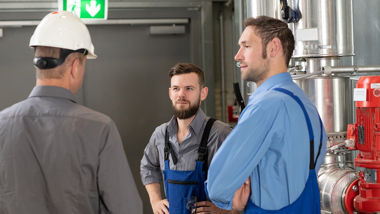 drei Männer stehen in einer Werkhalle, einer hat einen weißen Helm auf und steht mit Rücken ins Bild, er informiert die anderen beiden die in blauer Arebtiskleidung zuhören