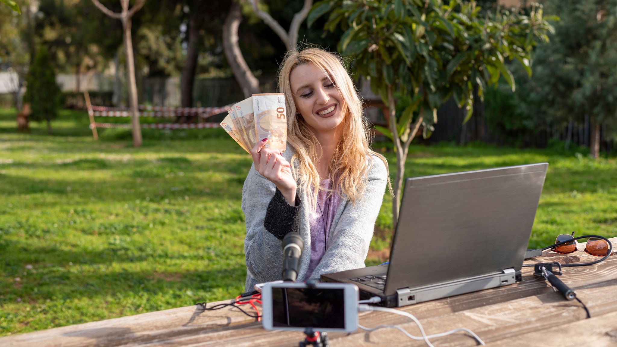Person sitzt mit Laptop und Mikrofon im Freien an einem Tisch und hält mehrere Euro-Banknoten in die Kamera.