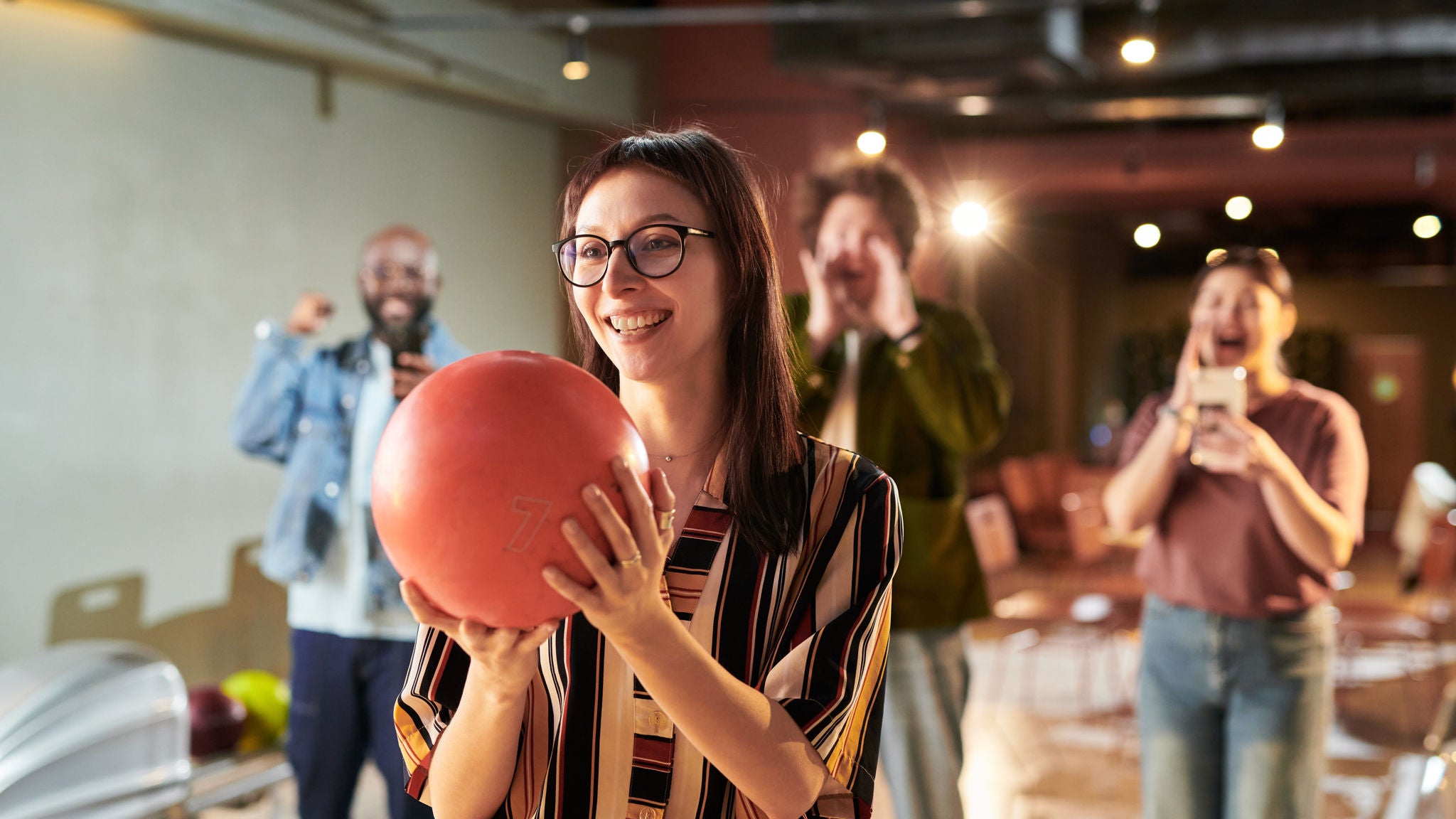 Frau mit Bowling-Kugel wird von Kolleg:innen im Hintergrund bejubelt.