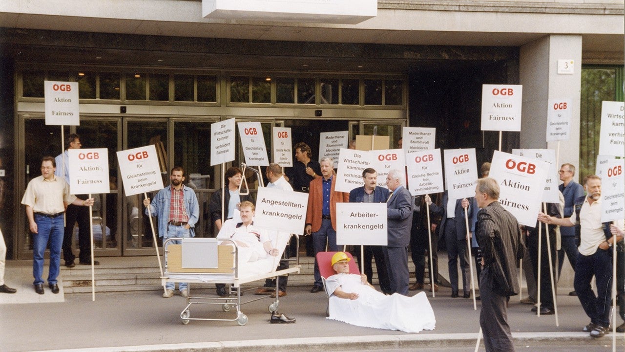 Vor den gläsernen  Türen einer Wirtschaftskammer (1999)  stehen Männer mit Plakaten mit ÖGB-Logo, Aufschrift: Aktion Fairness und Forderungen nach der Angleichung der ArbeiterInnenrechte an jene der Angestellten. In einem Krankenbett liegt ein "Angestellter" in einer roten Liege am Boden ein Arbeiter mit gelben Helm am Kopf.