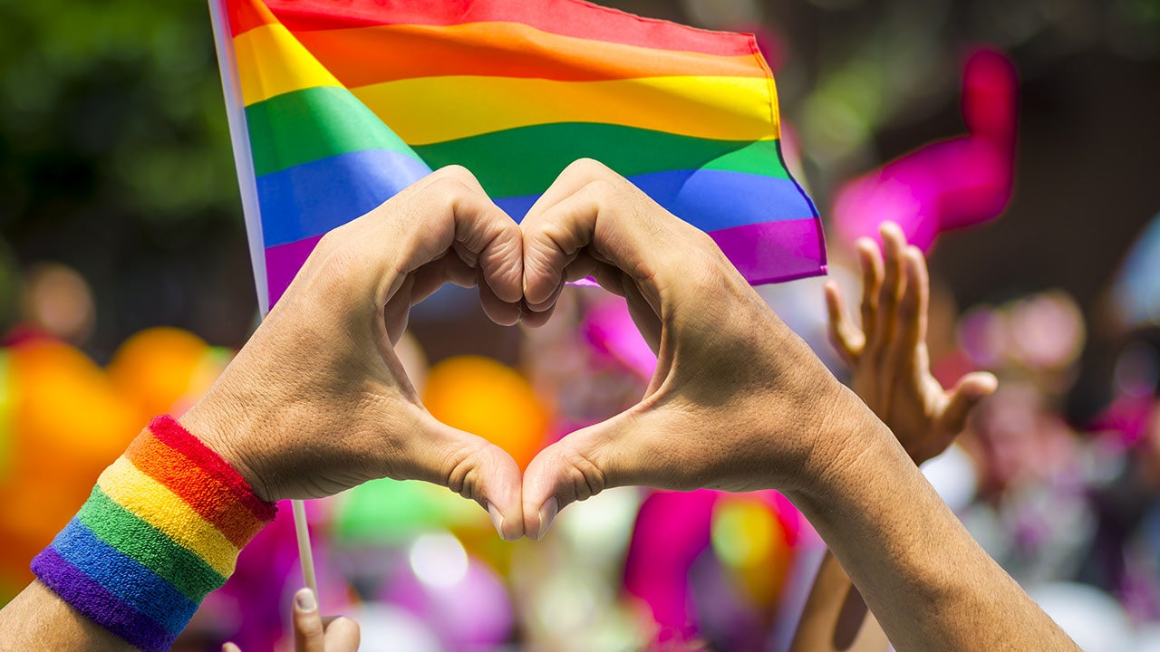 pride_haende_Herz eine Menschenmenge, im Vordergrund zwei Haende die eine Herzform bilden, im HIntergrund eine Pridefahne