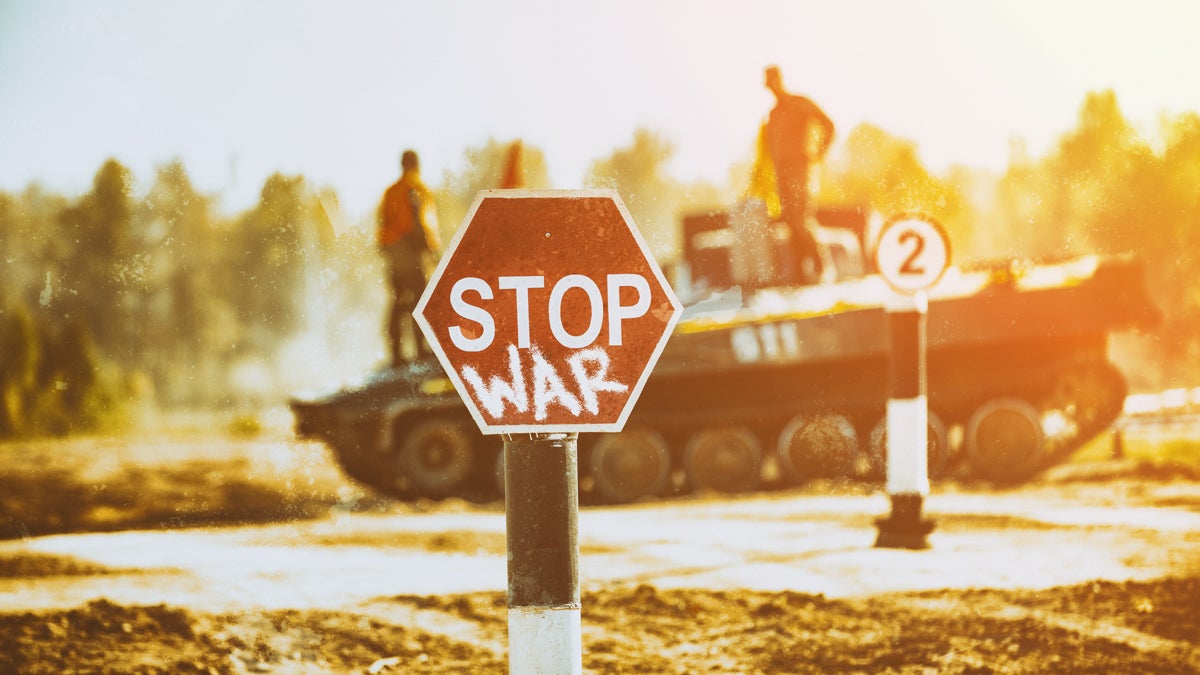 Im Hintergrund unscharf ein Panzer mit SoldatInnen, im Vordergrund ein "Stop"-Verkehrszeichen auf das "WAR" gesprayt wurde und nun die Botschaft "STOP WAR" trägt.