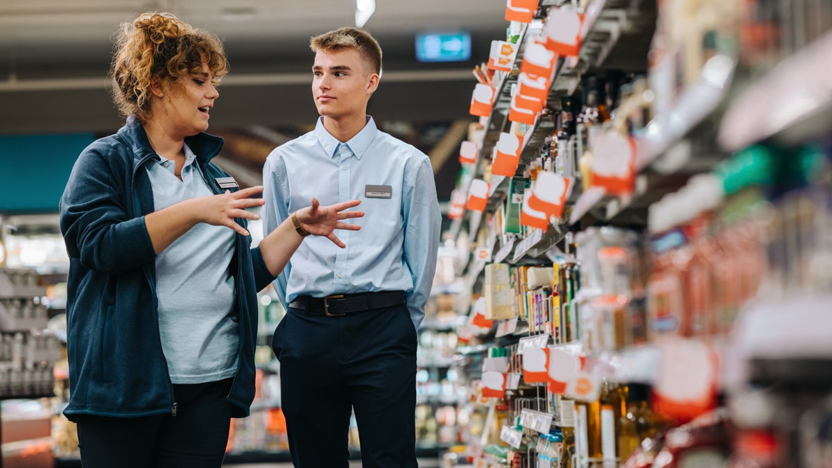 Supermarkt Managerin erklärt Lehrling etwas während sie beide vor einem Regal stehen. Das Regal und der Hintergrund sind unscharf, Produkte sind nicht zu erkennen.