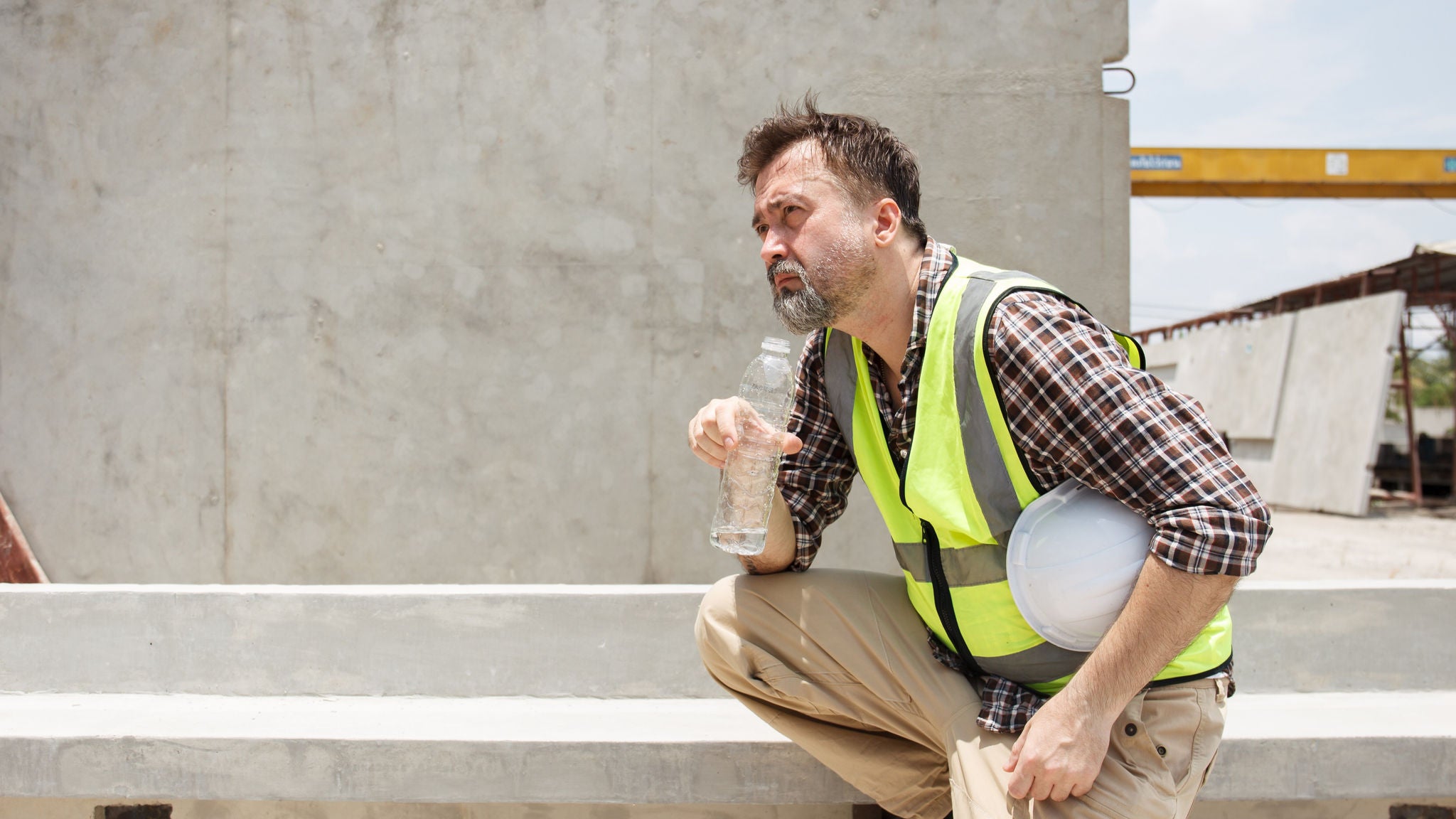 Bauarbeiter mit Warnweste und Schutzhelm hält auf einer Baustelle eine Wasserflasche in der Hand.