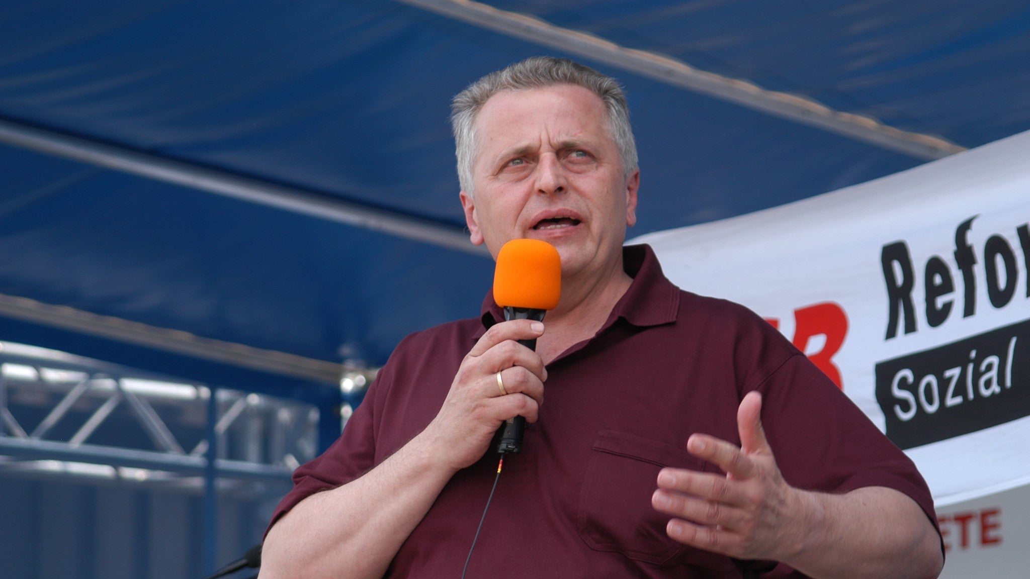 Rudolf Hundstorfer (rotes Poloshirt) spricht in Mikrofon. Dahinter Ausschnitte von Bannern