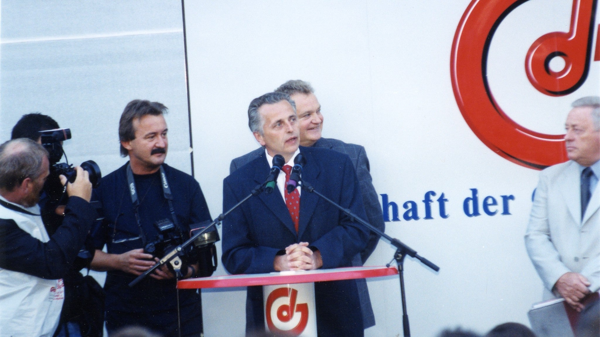 Rudolf Hundstorfer (blauer Anzug, rote Krawatte) spricht in zwei Mikrofone. Logos der Gewerkschaft der Gemeindebediensteten