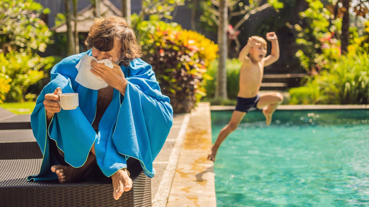 Mann sitzt mit einer Teetasse, in eine Decke eingewickelt am Pool und schnäuzt sich. Im Hintergrund springt ein Kind freudig in den Pool.