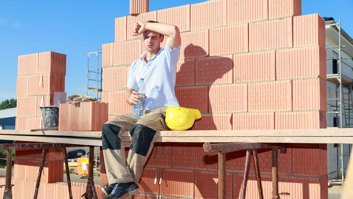 Bauarbeiter sitzt bei prallem Sonnenschein auf der Baustelle und wischt sich den Schweiß von der Stirn. Er hält eine Wasserflasche und hat seinen gelben Schutzhelm abgenommen.