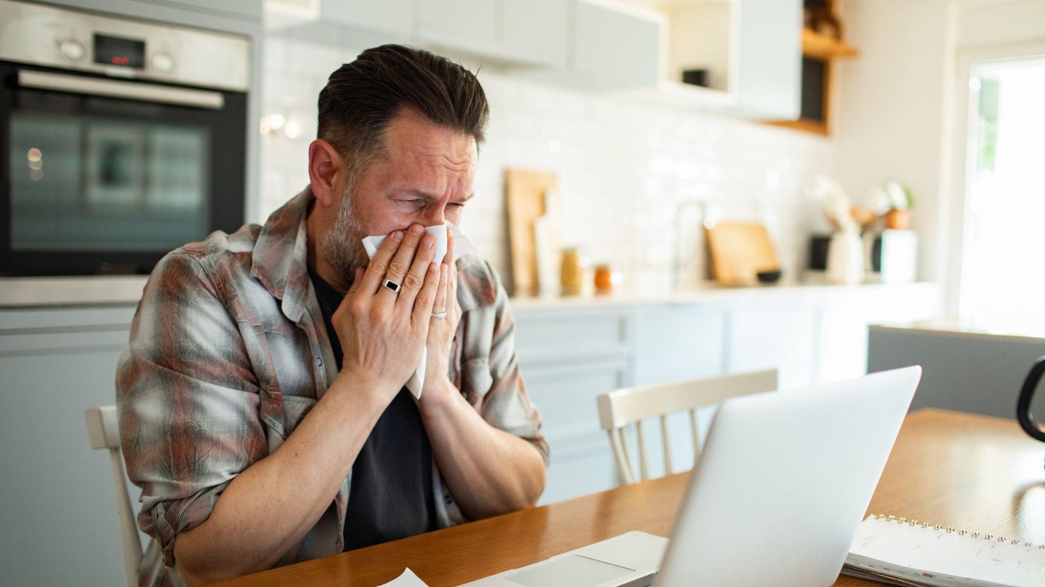 Ein Mann sitzt am Esstisch vor seinem Laptop und schnäuzt sich.