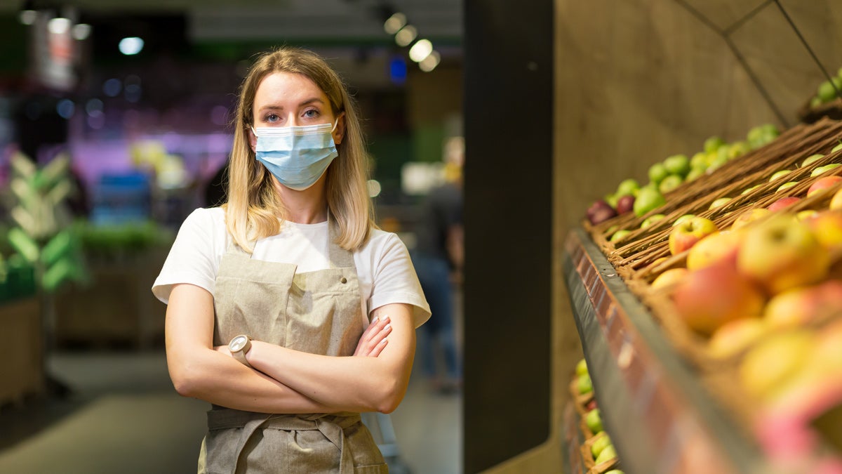 Supermarkterkäuferin steht mit verschränkten Armen in der Obstabteilung und schaut böse in die Kamera.