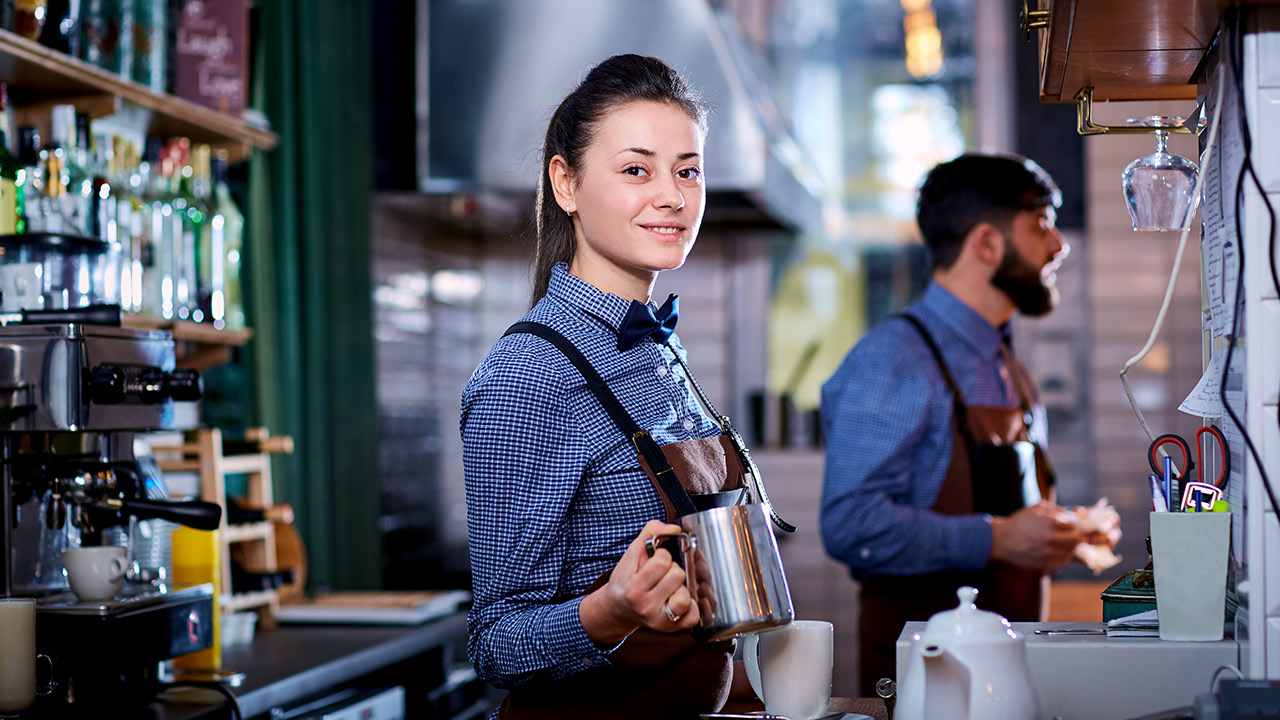junge Frau im Gastro-Service, lächelt, hinter der Theke, schaut in die Kamera, mit Milchschaum-Kanne in der Hand, im Hintergrund ein Mann der gerade eine Bestellung aufnimmt