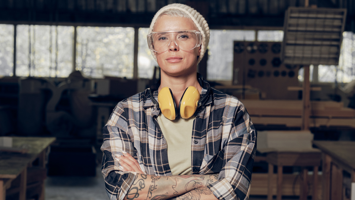 Portrait einer jungen Frau in einer Holzwerkstatt mit Sicherheitsbrille und Gehörschutz.