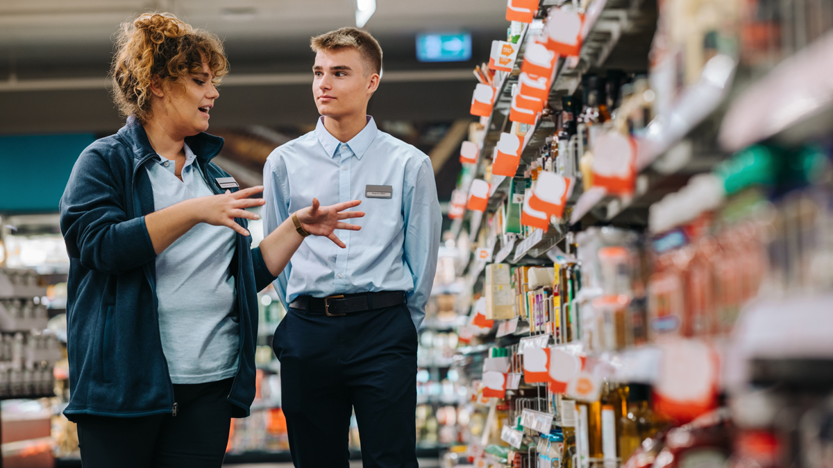 Ferialjob/Lehrling Supermarkt Managerin erklärt Lehrling etwas während sie beide vor einem Regal stehen. Das Regal und der Hintergrund sind unscharf, Produkte sind nicht zu erkennen.