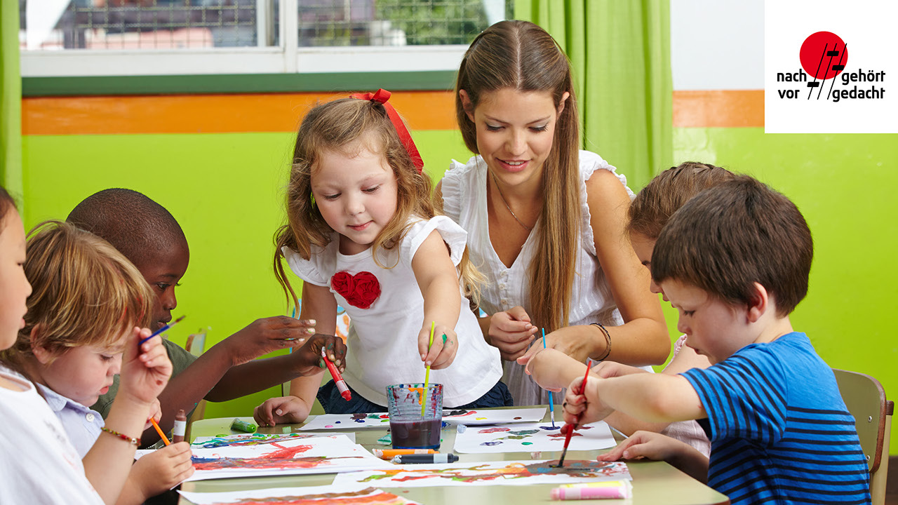 eine Elementarpaedagogin sitzt mit eine Gruppe von Kleinkindern an einem Tisch und hilft ihnen beim malen und Zeichnen
