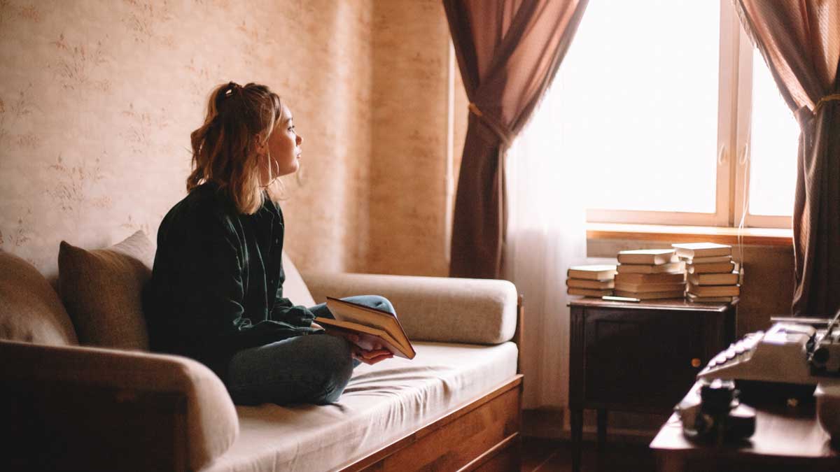 Junge Frau sitzt, mit einem Buch in der Hand, auf der Couch und schaut besorgt aus dem Fenster