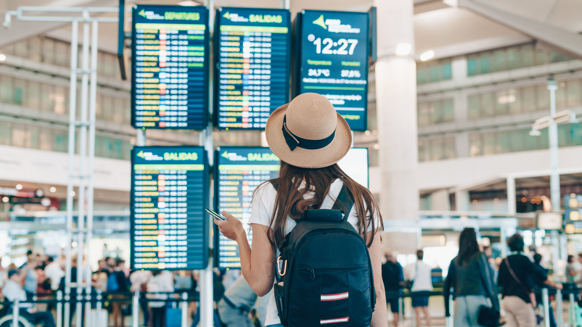 Frau steht  mit Gepäck am Flughafen und betrachtet die Abflug-Informations-Tafeln.