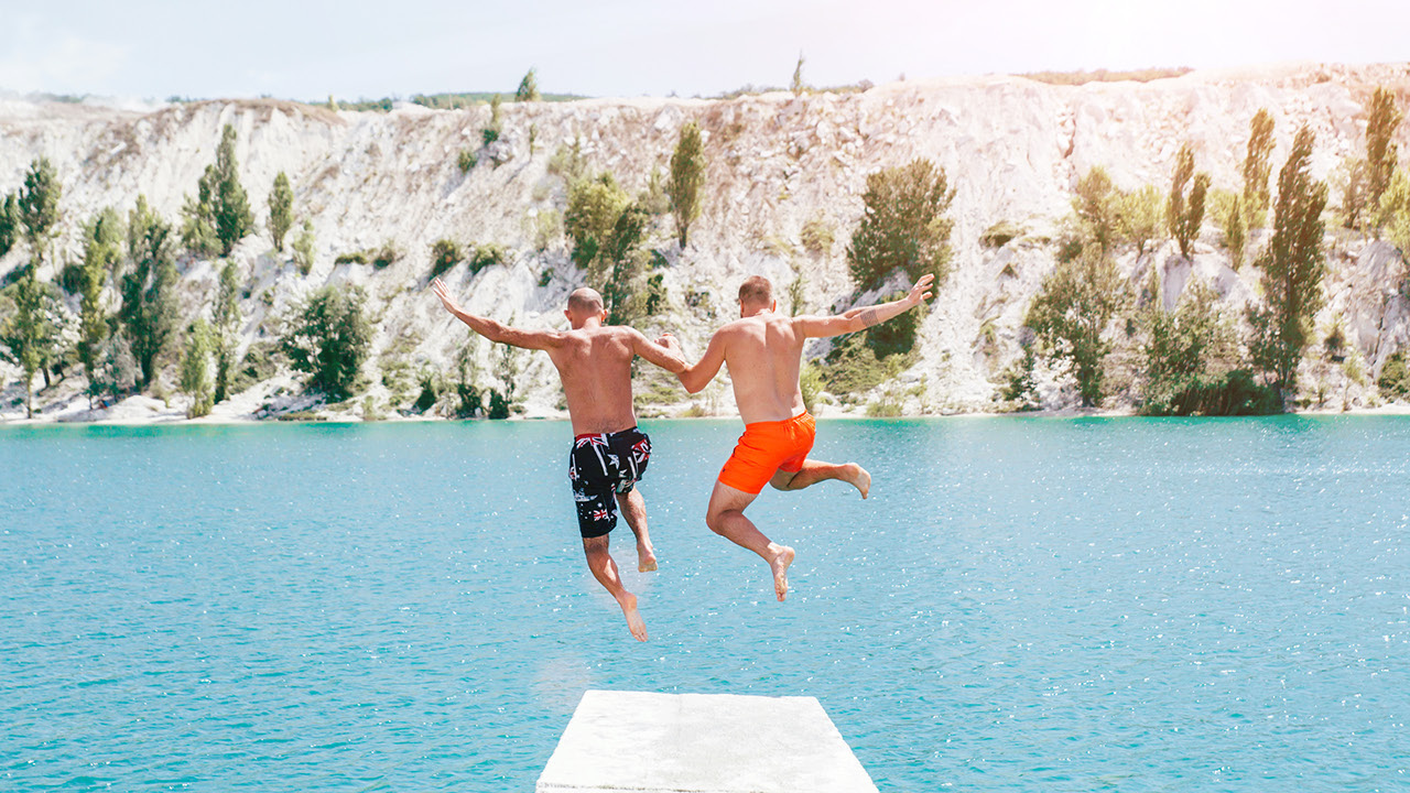 zwei Männer halten sich an den Händen und springen ins Wasser. Türkises Wasser und Felsen am anderen Ufer. Urlaubsstimmung