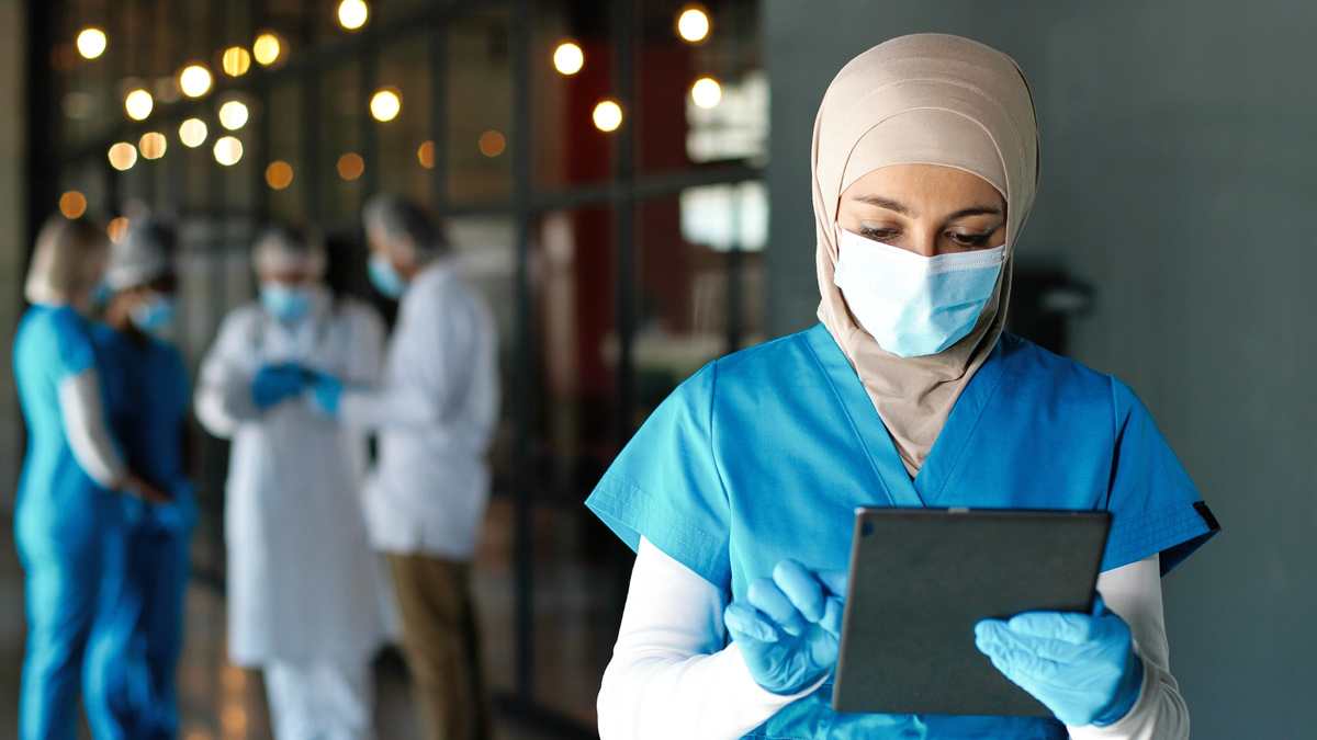Krankenpflegerin mit Kopftuch, Maske und Handschuhen arbeitet auf einem Tablet.