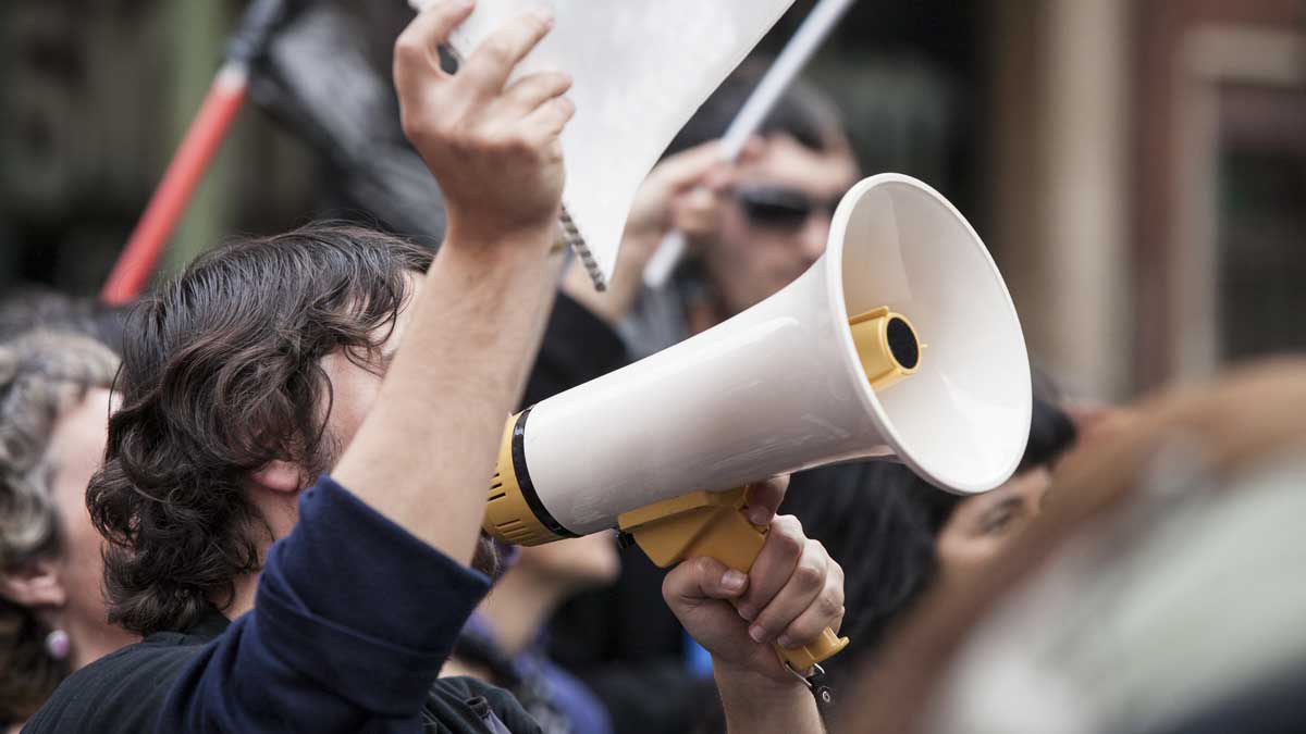 Foto von einem Protest. Im Vordergrund ein Mann mit Megafon der ein Schild hochhält.