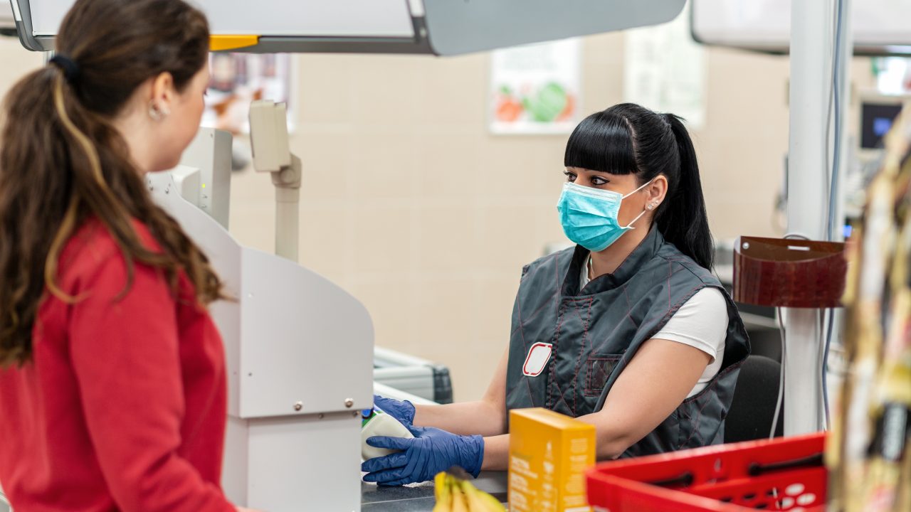 A young woman in a medical mask and gloves, working at the checkout in a supermarket. In the foreground is a customer in a blur. Concept of coronovirus, protection from infection and industrial crisis.